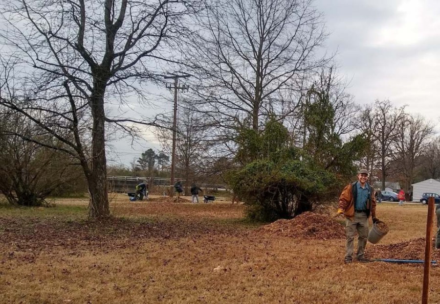 Foreground man smiling holding a bucket in a park. In the background, volunteers are spreading mulch around saplings.