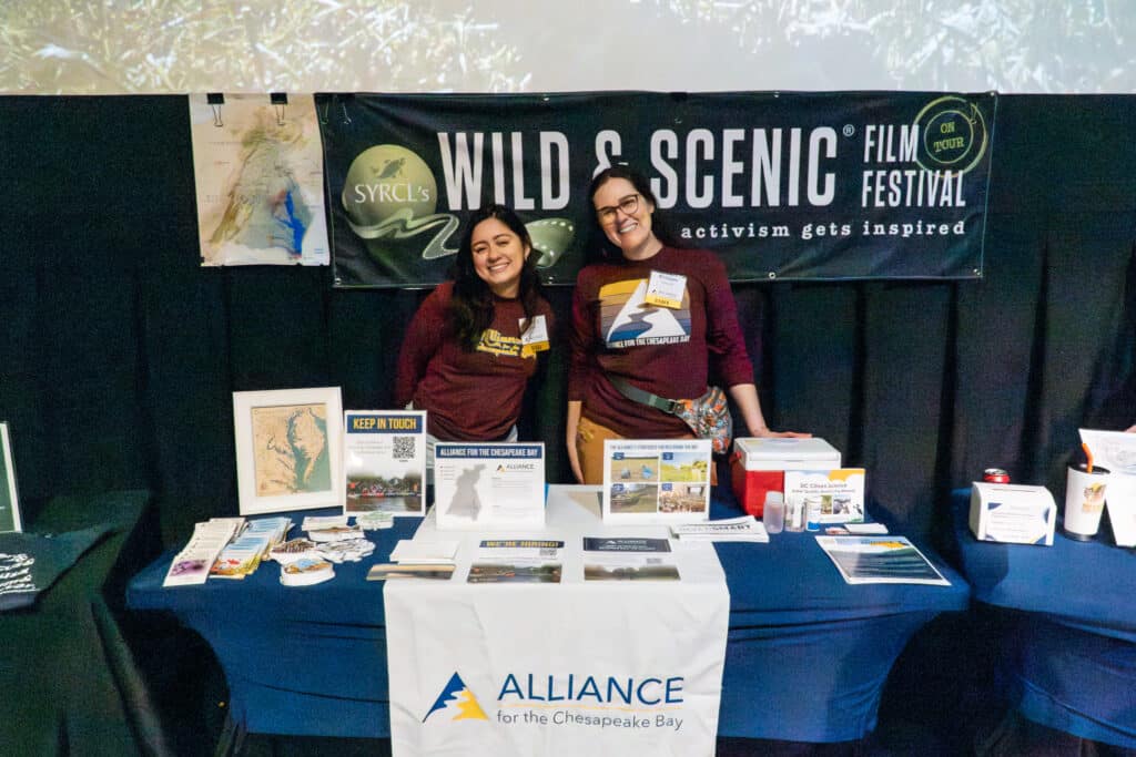 Two Alliance staff members smiling as they stand behind a table set up with Alliance logos and educational materials.