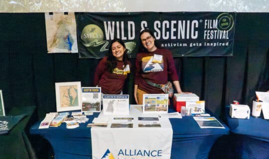 Two Alliance staff members smiling as they stand behind a table set up with Alliance logos and educational materials.