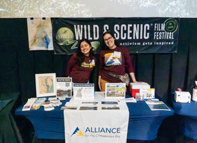 Two Alliance staff members smiling as they stand behind a table set up with Alliance logos and educational materials.