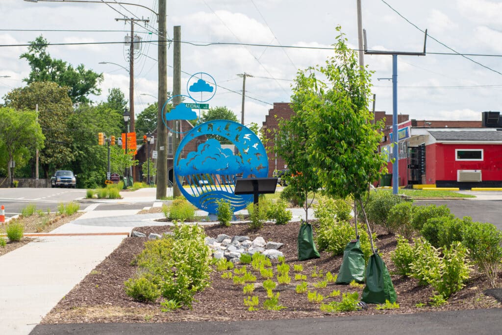 A bright blue sculpture with clouds and birds sits amongst plants in a newly planted rain garden.