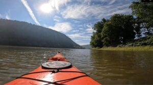 The front of a kayak looking toward a hill, with the sun shining directly above it.