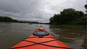 The view from inside of a kayak going down a river during a cloudy day.