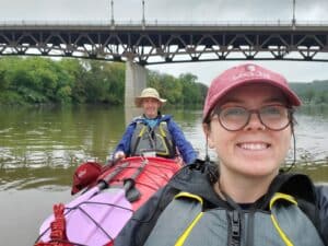 Two people floating down a river in kayaks, past a bridge.