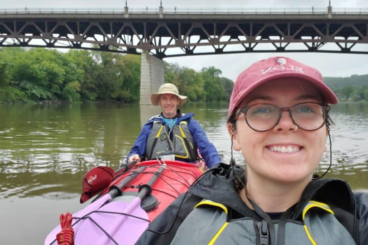 Two people floating down a river in kayaks, past a bridge.