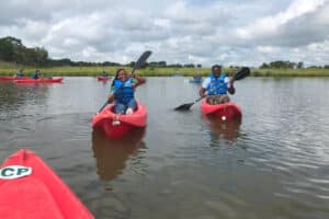 Two students smiling and kayaking in the foreground with many other students kayaking in the background