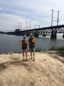 Two people holding up balloons on the shore of a river, with a large bridge in the background