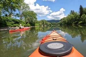 The view from inside of a kayak on a rive, with another kayaker to the left. Green trees in the foreground and sunny blue skies in the background.