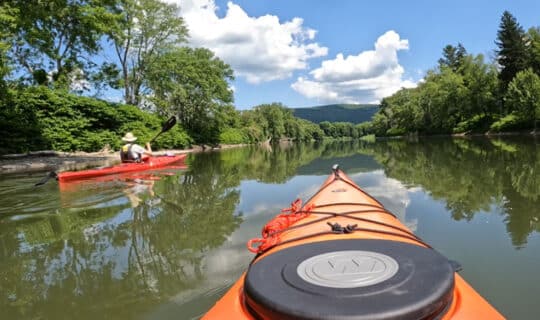 The view from inside of a kayak on a rive, with another kayaker to the left. Green trees in the foreground and sunny blue skies in the background.