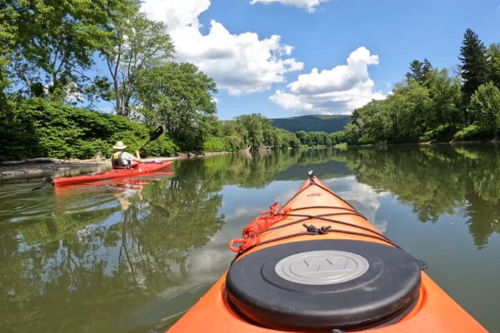 The view from inside of a kayak on a rive, with another kayaker to the left. Green trees in the foreground and sunny blue skies in the background.
