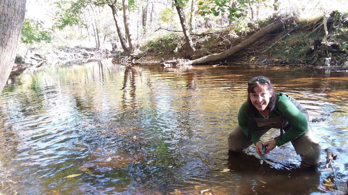 A person crouching in a shallow creek, smiling at the camera