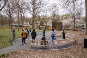 Thirteen people stand in a circle in a park.