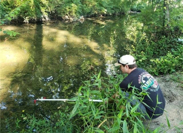 A person crouched next to a creek placing a pole into the water