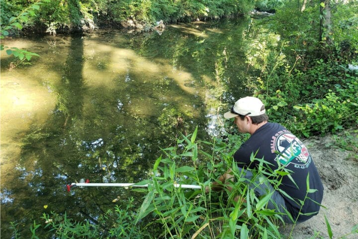 A person crouched next to a creek placing a pole into the water