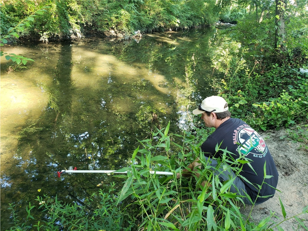 A person crouched next to a creek placing a pole into the water