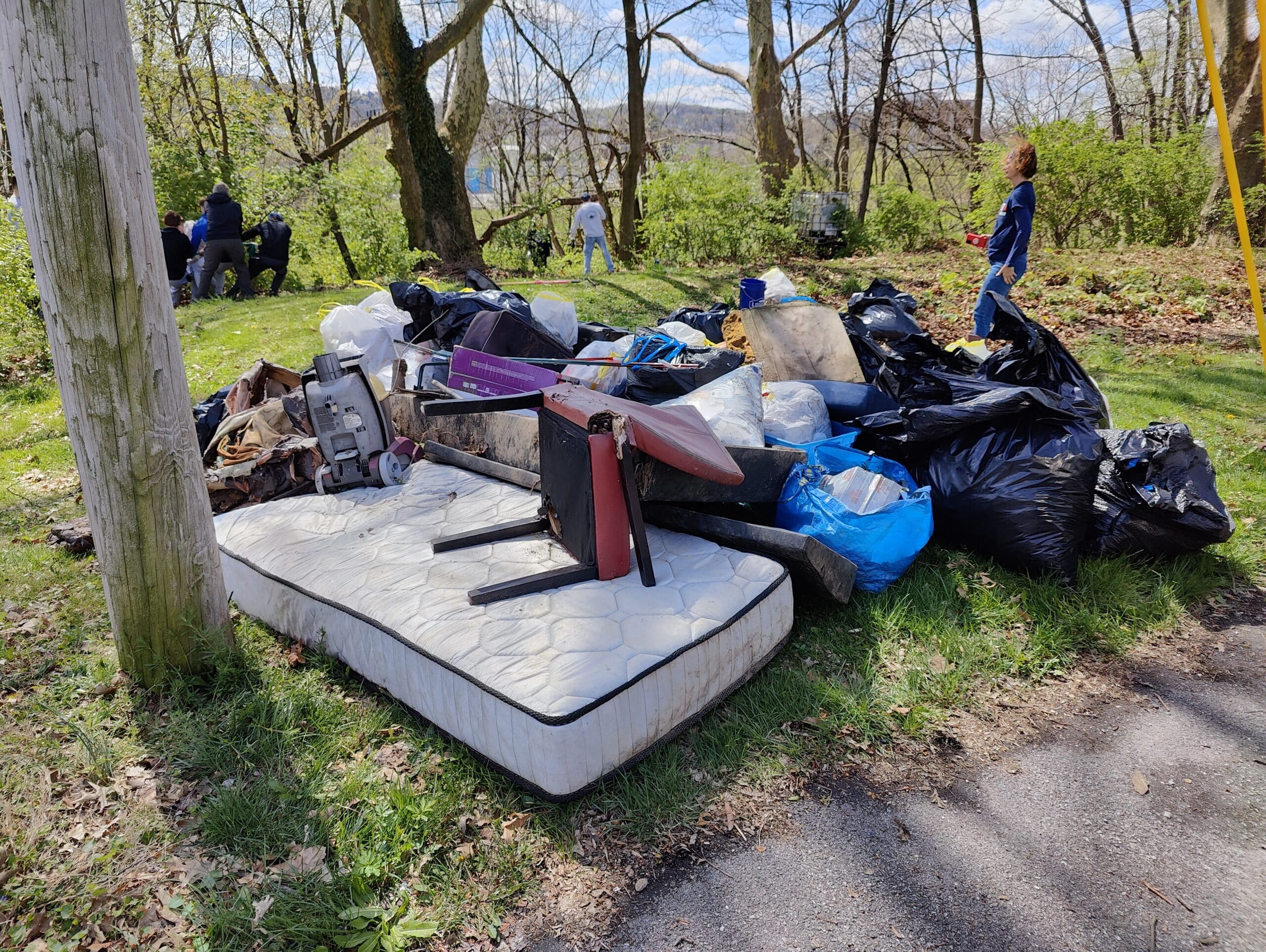 A mattress, a chair, and a mound of trash bags sits on the edge of parking lot in the grass.