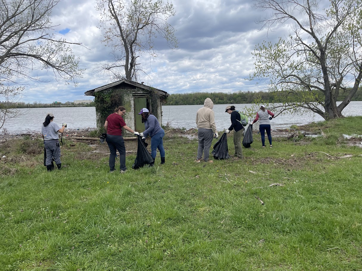 A group of volunteers holding trash bags and trash pickers collect trash in front of the Potomac River.