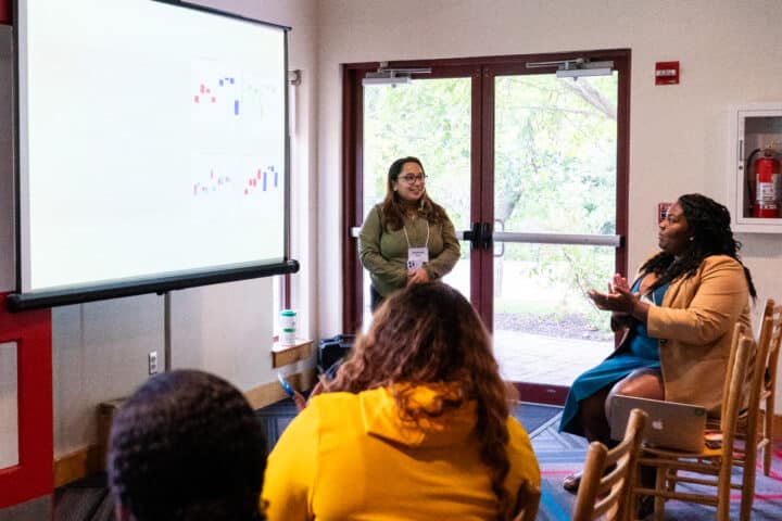 A person standing in front of a projector screen presenting to a small group of individuals seated in a classroom.