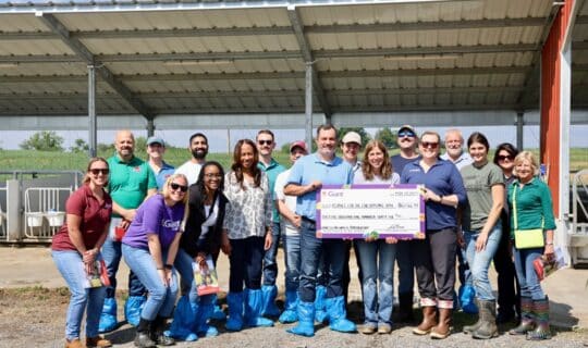 A group of people stand around a large check on a farm property.