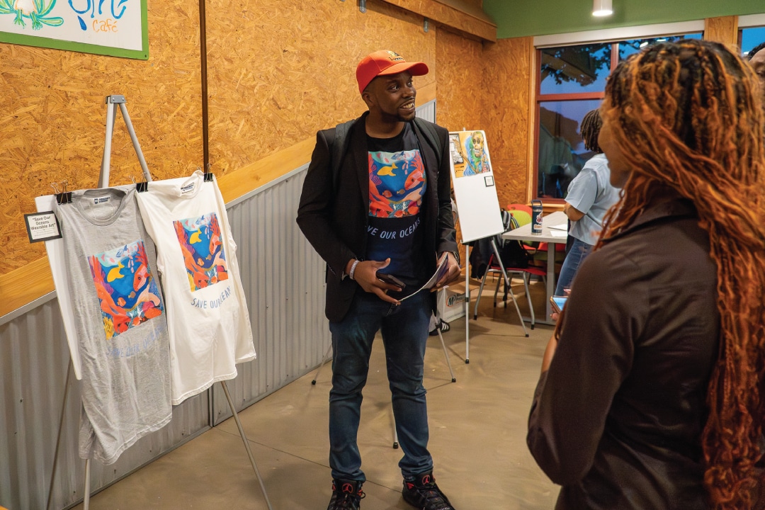 a person speaks with others while standing next to displayed shirts