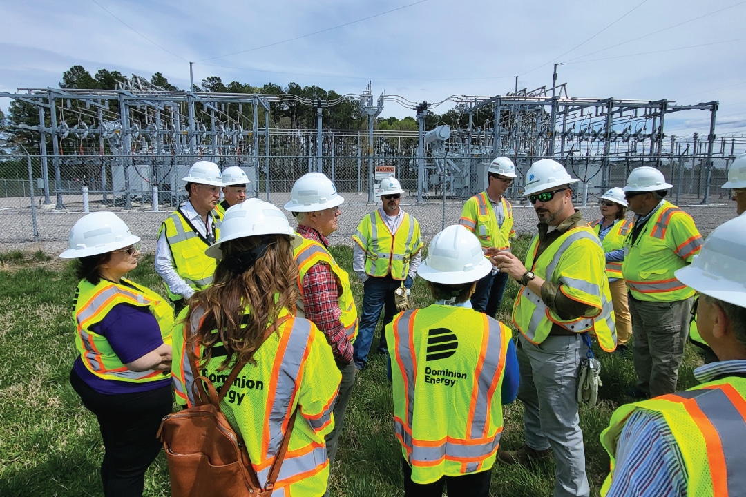 a group of people listen to a speaker while wearing hi-visibility vests and hardhats