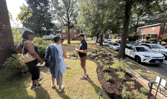 An Alliance team member stands in front of a rain garden on a residential property while talking to two community members.